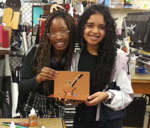Two people proudly holding up a paper-cut project. They are surrounded by classroom tools.